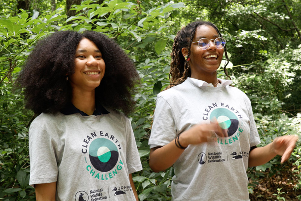 Two teenagers with matching "Clean Earth Challenge" tshirts smile as they walk through a forest