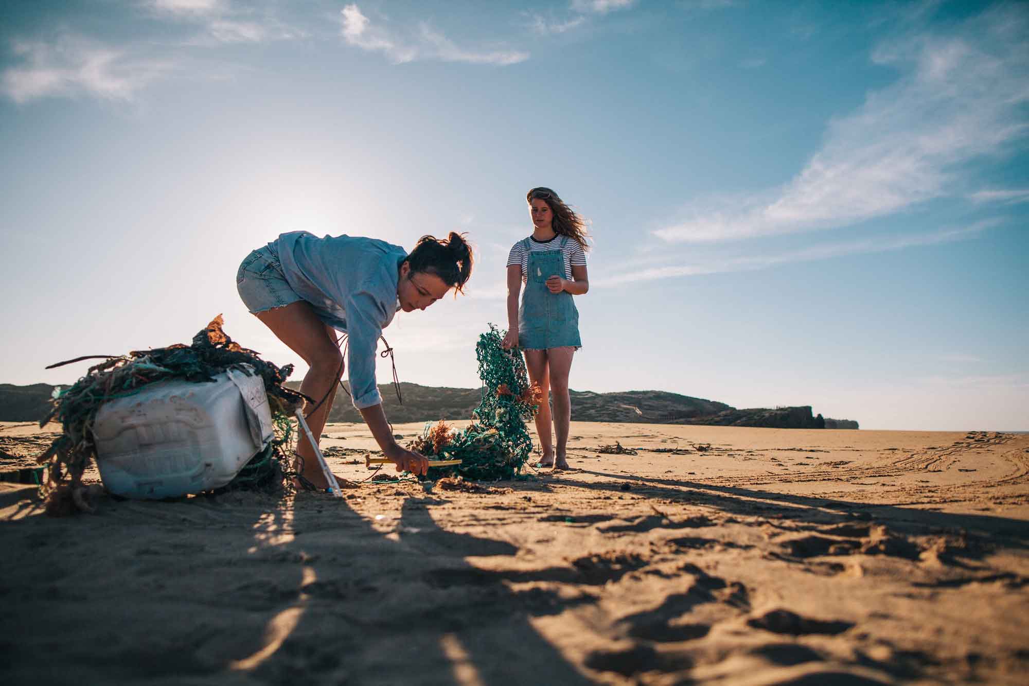 Two women picking up plastic litter and debris on a sandy beach during sunset.