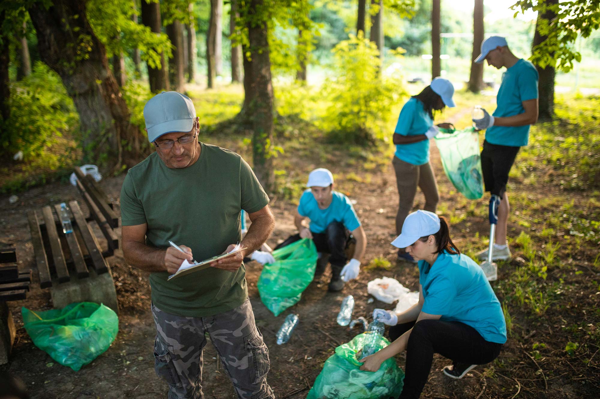 An older man marks an a clipboard as team members behind him continue to clean up trash in a park.